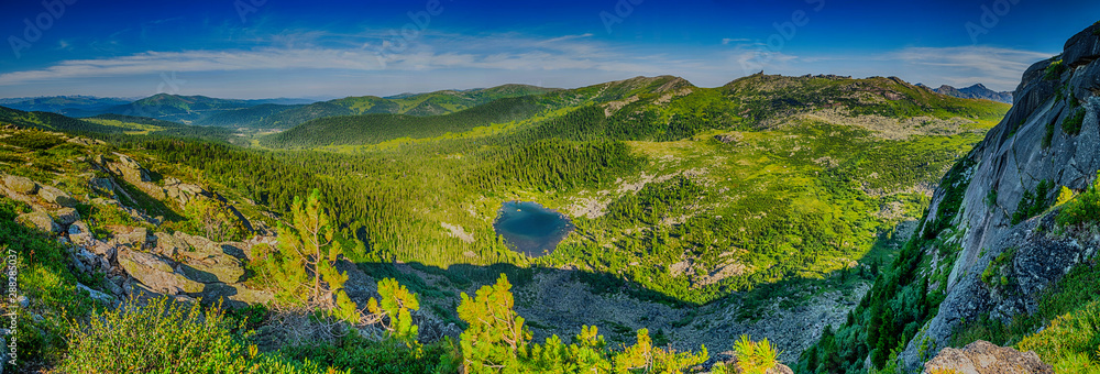 Panoramic view from the cliff on beautifull valley with small mountain lake, green forest and sayan mountain range in Ergaki nature park in Krasnoyarsk region, Russia