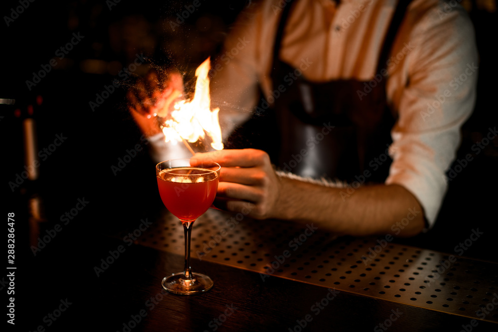 Professional bartender serving a cocktail in the glass spraying on the burning match with a lemon juice