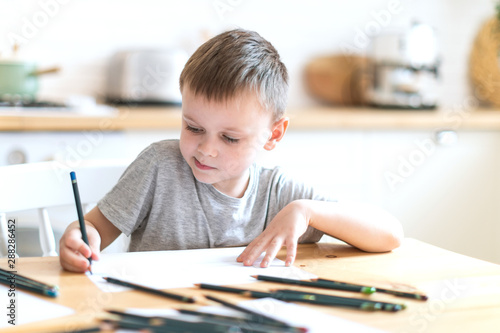 Little boy sits at a table in a bright kitchen and draws with pencils