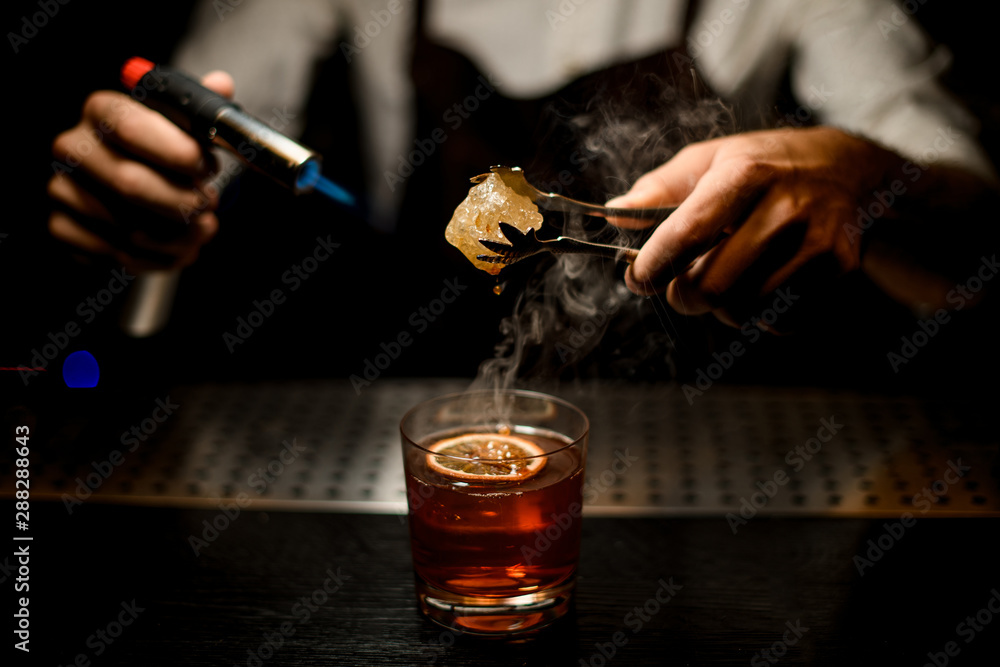 Professional male bartender serving a brown cocktail melting caramel with a burner above the lemon slice in the dark