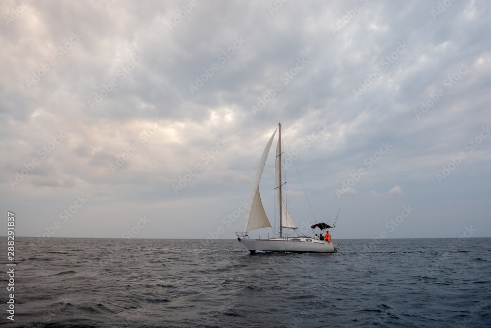 White sail boat on the grey calm water of sea or ocean during cloudy, windy and grey day with yacht team on board 