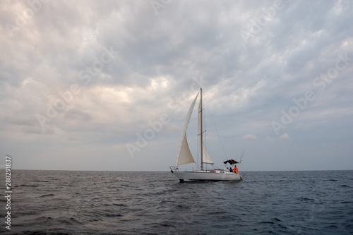White sail boat on the grey calm water of sea or ocean during cloudy, windy and grey day with yacht team on board 