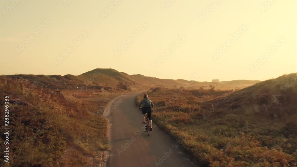 Tracking aerial shot of a young woman cyclist riding a bike on a bike path among sand dunes at golden sunrise in the unique landscape of the Texel island in The Netherlands