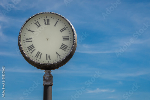 Abstract old fashioned street clock with Roman numerals without arrows on blue sky background. Concept of stopped time