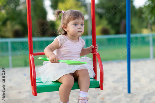 Funny little child, adorable preschooler girl in pretty dress having fun on a swing in the park on summer day