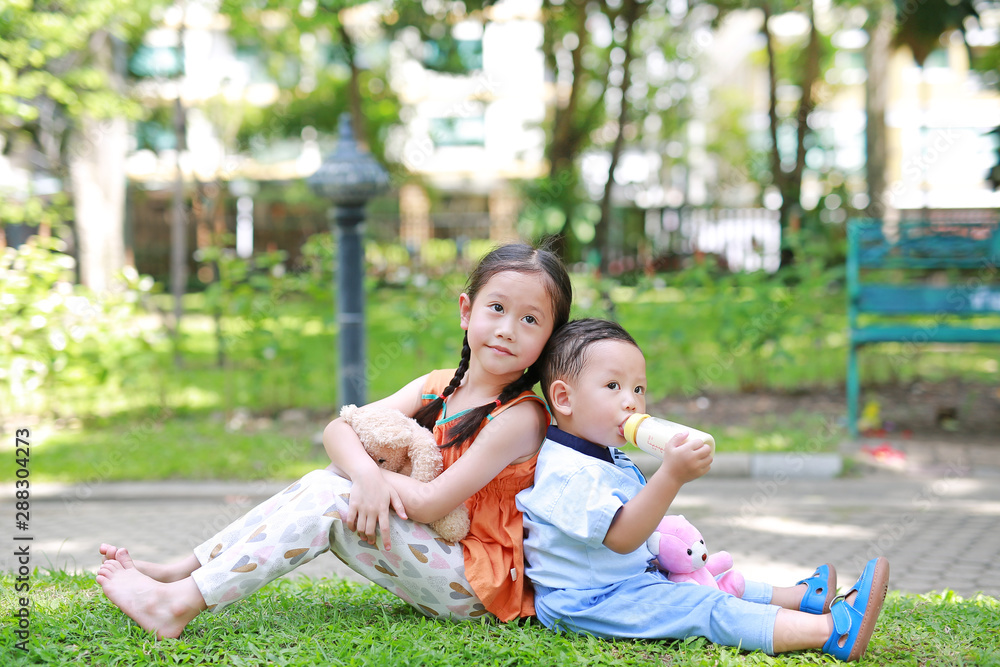Happy little Asian sister and her younger brother sit back and lean back together in the green garden. Child girl cuddle teddy bear doll and boy Suck milk from bottle. Happy little Asian sister and her younger brother sit back and lean back together in the green garden. Child girl cuddle teddy bear doll and boy Suck milk from bottle.