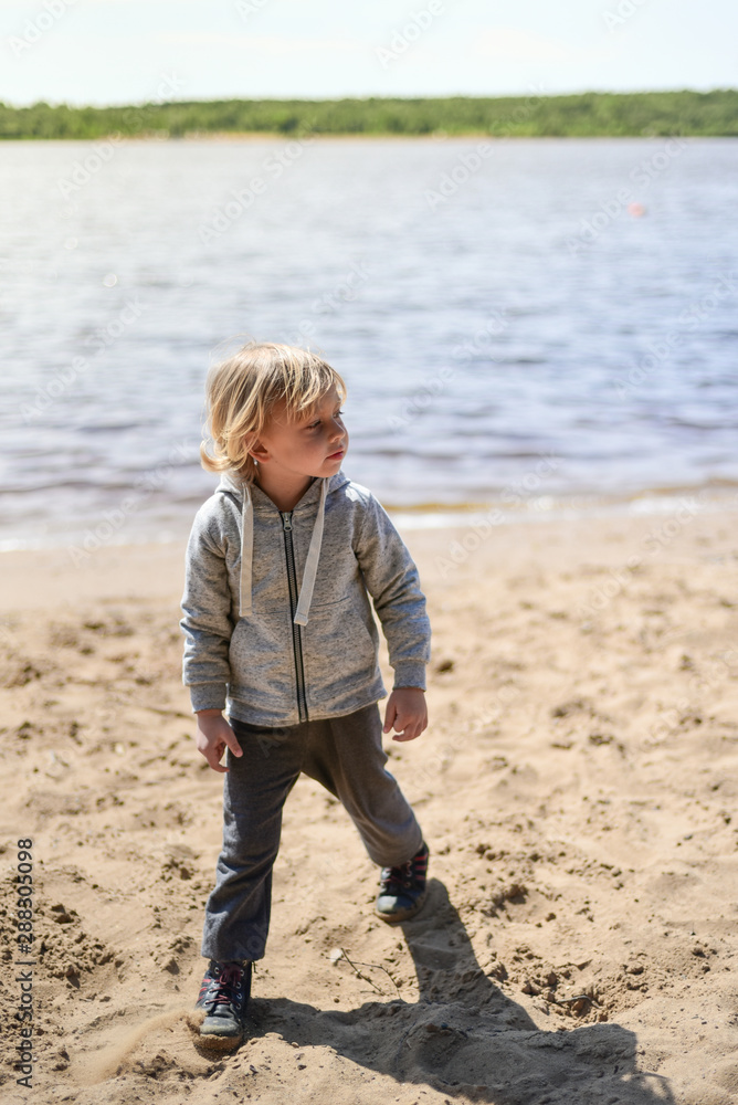 Blond kid walking on the beach in light clothes. Warm summer day. A lonely boy on the river Bank. Emotions. Walk with children. Blond kid walking on the beach in light clothes. Warm summer day. A lonely boy on the river Bank. Emotions. Walk with children.