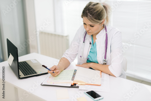 Beautiful young woman doctor sitting at the desk and writing against the background of a window. There s a laptop on the table