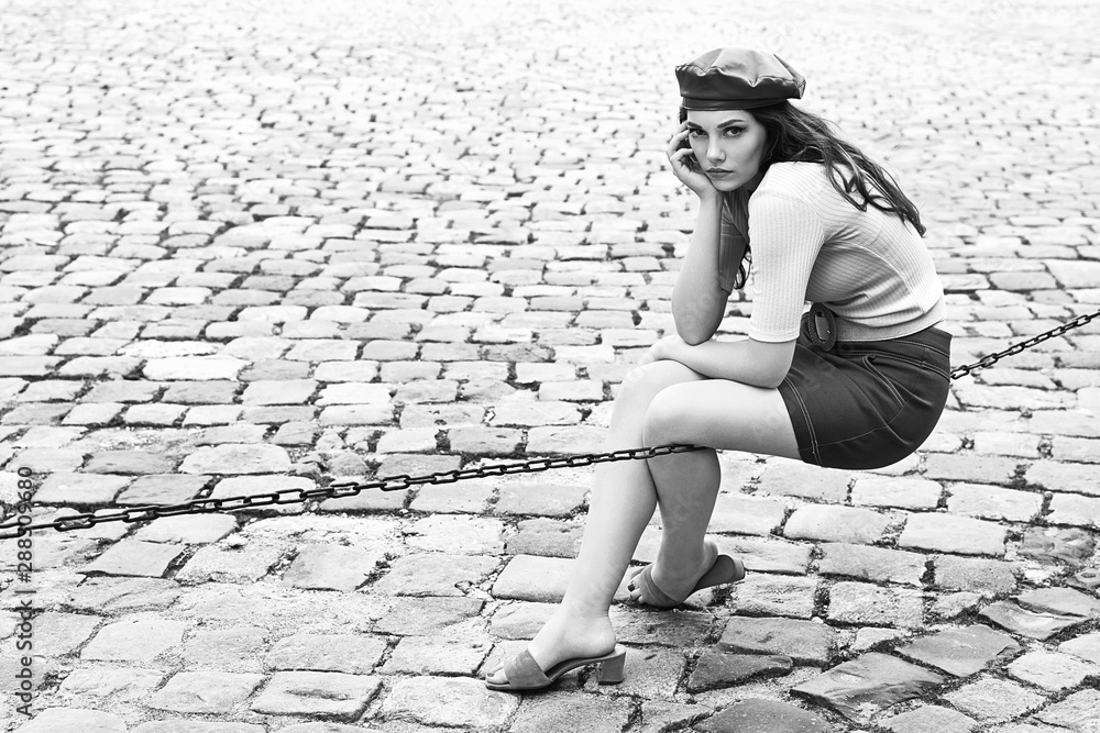 Young beautiful brunette emotional girl dressed in retro vintage style in the old european city sits of a chain