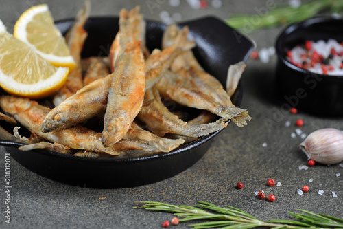 Fried smelt fish with lemon slices in a black pan. Dark background. Garlic, rosemary, spices and lemon slices complete the composition.