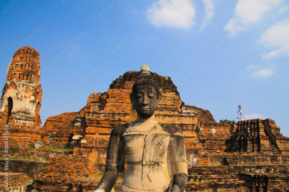 View on isolated old Buddha statue with raised left hand against blue sky and ancient red brick temple ruin (Focus on Buddha statue) - Ayutthaya, Thailand