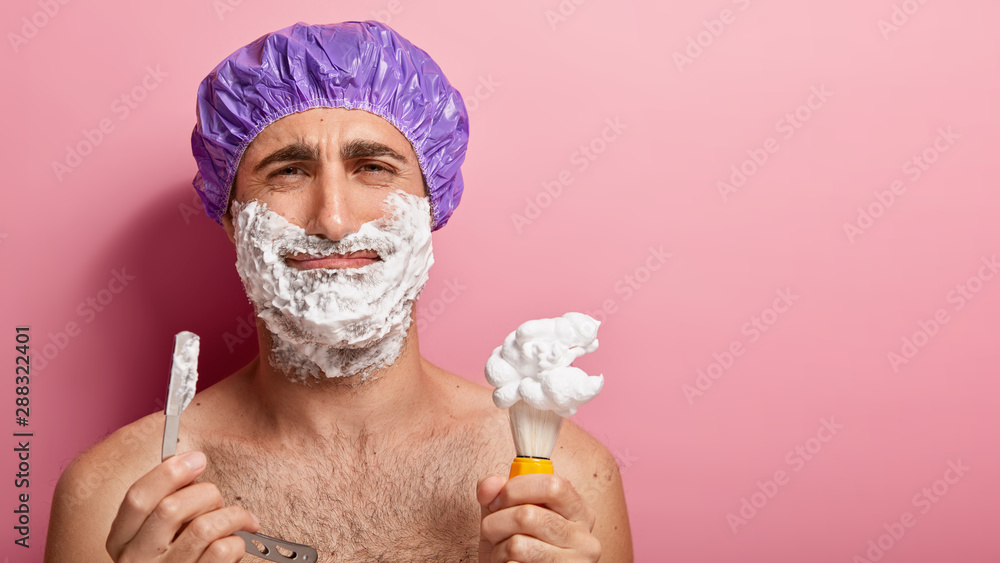 Unhappy man applies shaving gel on cheeks, fed up of daily routine, shaves stubble, holds blade
