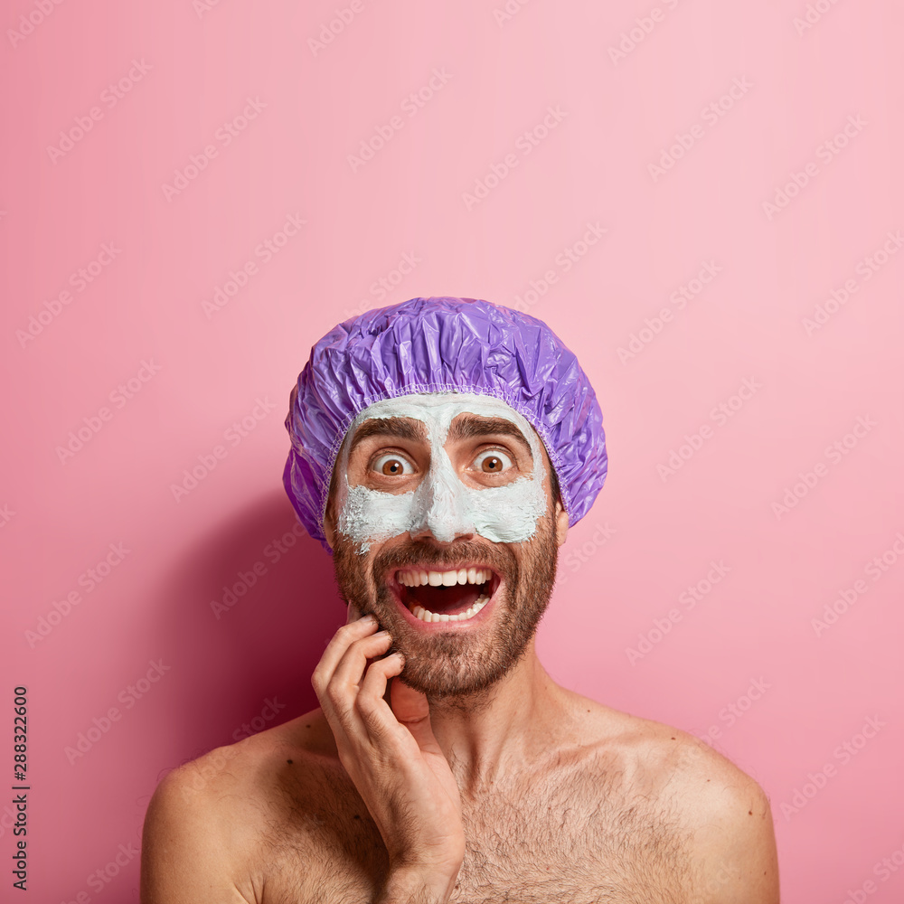 Worried young man applies white mud mask on face, feels refreshment and rejuvenation, wears bathcap, stands shirtless against pink background, copy space above. Face care, softness and purity Worried young man applies white mud mask on face, feels refreshment and rejuvenation, wears bathcap, stands shirtless against pink background, copy space above. Face care, softness and purity