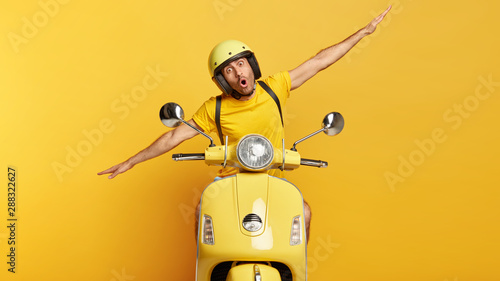 Impressed young male motorcyclist spreads hands sideways, drives motorbike, enjoys extreme drive, wears yellow t shirt, helmet, isolated over yellow background. Monochrome shot. Transport and people