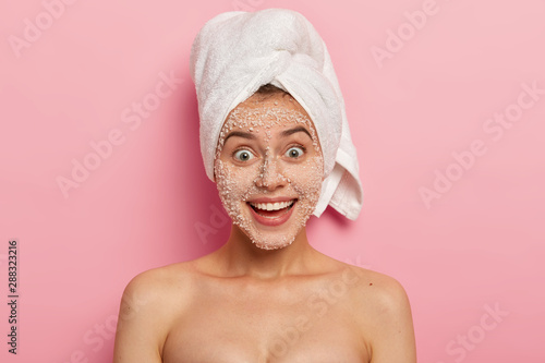 Facial treatment. Lovely happy woman with charming smile, removes toxins and blackheads on face, applies natural scrub of white sea salt granules, pulls out clogs, has naked well cared body.