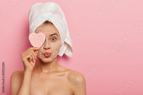 Reducing pores and cleansing concept. Attractive female applies sea salt mask on face, has luxurious feelings from beauty treatments, covers eye with heart shaped sponge, pampers complexion. photo
