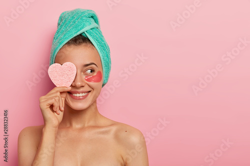 Cheerful young female model has pleasant smile, covers eye with cosmetic sponge, enjoys all benefits of patches, reduces wrinkles, wears wrapped towel on head, has skincare routine after awakening photo