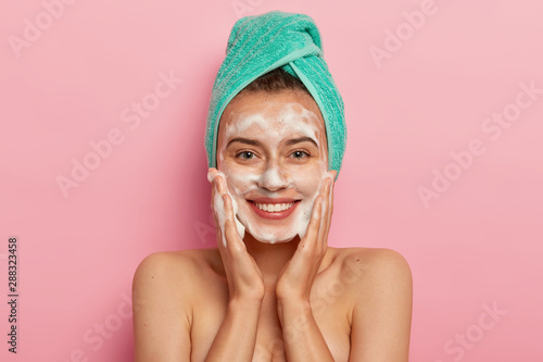 Positive young female has toothy smile, has perfect teeth, pats skin with liquid sanitary soap, washes with foaming gel, wakes up in morning for having beauty routine, isolated on pink background