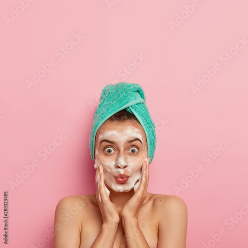Skincare routine and hygiene concept. Photo of lovely woman with widely opened eyes, keeps lips folded, massages cheeks and applies foaming soap, poses against pink background, copy space above