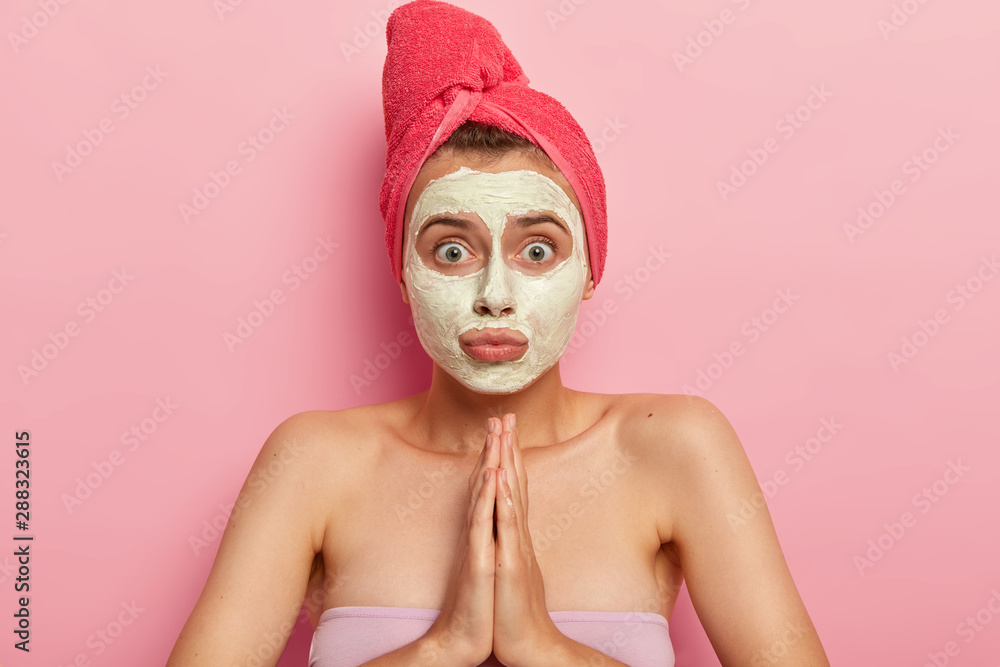 Photo of surprised young woman keeps palms together in praying gesture, has helpess puzzled face expression, wears nourishing facial mask for reducing wrinkles, uses homemade cosmetic ingredients Photo of surprised young woman keeps palms together in praying gesture, has helpess puzzled face expression, wears nourishing facial mask for reducing wrinkles, uses homemade cosmetic ingredients