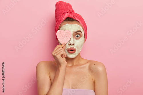 Facial treatment and spa concept. Astonished young woman applies clay mask, shocked with quick effective result, keeps cosmetic sponge on eye, stands naked against pink background, heals skin