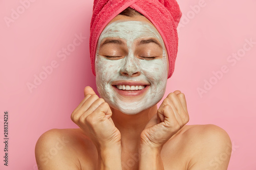 Close up portrait of happy female model with facial clay mask on face, clenches fists, receives good news, keeps eyes closed, wears pink towel on head, stands bare shoulders has rejuvenation procedure