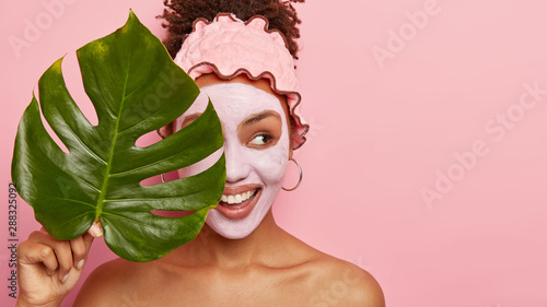 Cropped shot of happy young woman applies facial mud mask on face for removing wrinkless or fine lines, takes bath at bathroom, wears protective rosy headband, holds big plant leaf covers eyes with it photo