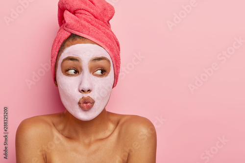 Indoor shot of good looking dark skinned model makes lips folded as if wanting to kiss someone, wears nourishing clay mask, wrapped in towel, looks aside on blank space, isolated on pink wall