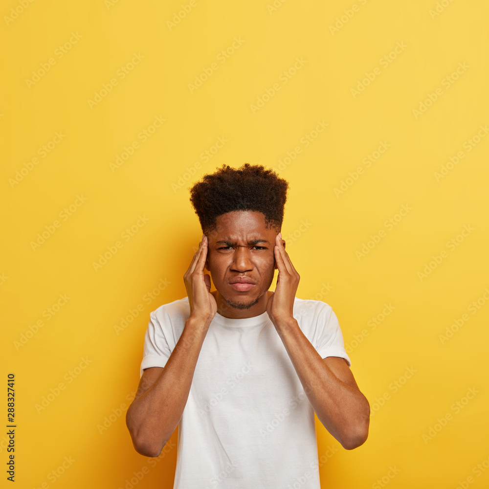 Vertical shot of dissatisfied dark skinned man student keeps hands on temples, suffers from terrible headache, wears white casual t shirt, stands against yellow background, copy space for your promo Vertical shot of dissatisfied dark skinned man student keeps hands on temples, suffers from terrible headache, wears white casual t shirt, stands against yellow background, copy space for your promo