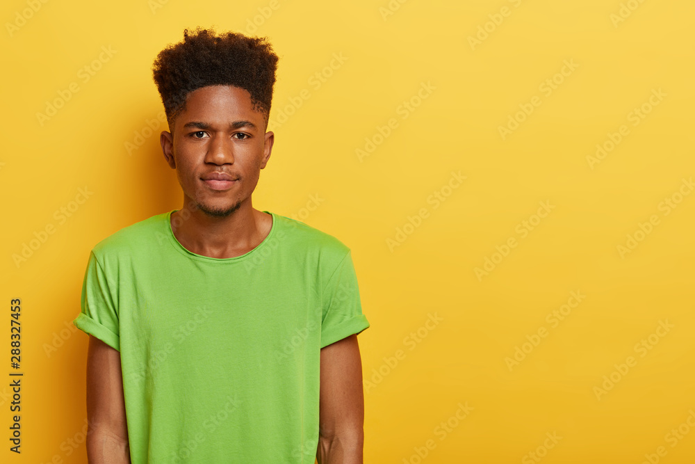 Photo of handsome teenage boy with dark skin, curly hairstyle, wears casual  green t shirt, looks with calm serious expression at camera, isolated over  yellow background, blank space on right side Stock