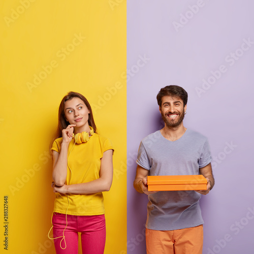 Vertical shot of thoughtful female stands in pensive pose, thinks something over, wears headphones around neck, wears yellow t shirt and pink pants, cheerful man holds cardboard box in hands