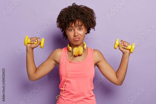 Photo of dissatisfied woman with curly bushy hair, busy doing biceps exercises, listens coach unwillingly, raises arms with dumbbells, wears casual pink vest, isolated over purple studio wall photo