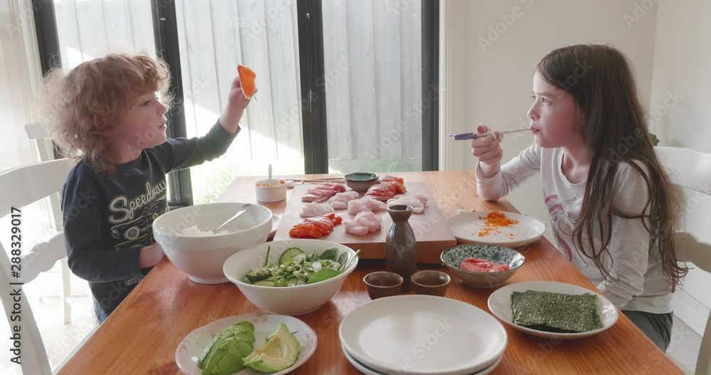 young boy and girl eat sashimi, raw fish, from a large serving of fish on a board on a table between them. There are also bowls of salad, rice and nori - seaweed