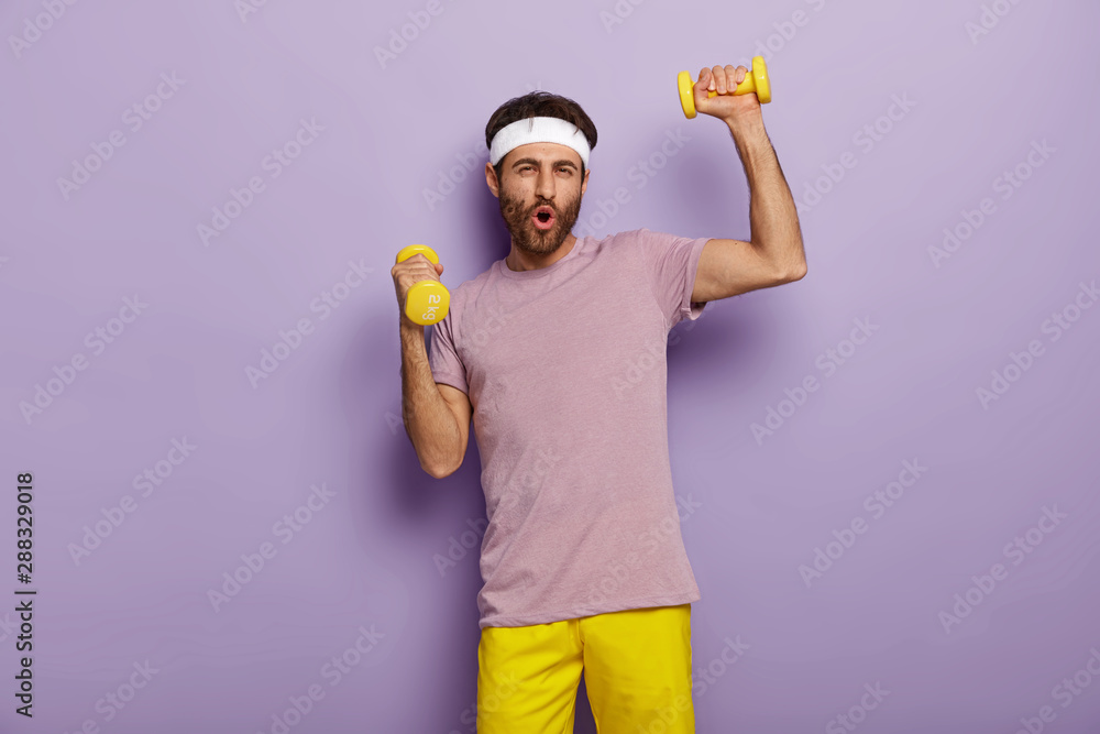 Funny man has fun, exercises with dumbbells, dressed in active wear, motivated for healthy lifestyle, has regular training in morning, isolated over purple background. Athletic guy holds weights Funny man has fun, exercises with dumbbells, dressed in active wear, motivated for healthy lifestyle, has regular training in morning, isolated over purple background. Athletic guy holds weights