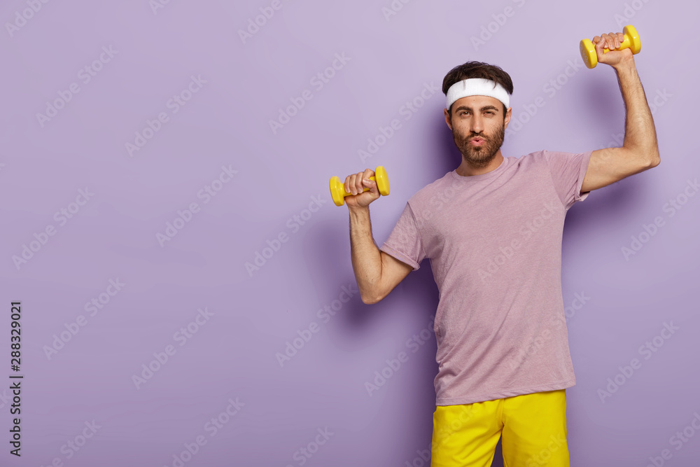 Horizontal shot of handsome unshaven man has workout in gym, trains biceps with sport instructor, wears active wear, white headband, stands against purple studio wall, copy space aside. Exercising Horizontal shot of handsome unshaven man has workout in gym, trains biceps with sport instructor, wears active wear, white headband, stands against purple studio wall, copy space aside. Exercising