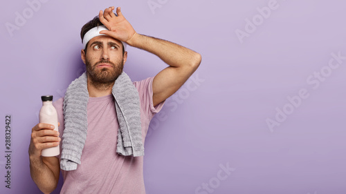 Exhausted athlete man feel thirsty and tired after hard cardio workout, wipes sweat on forehead, holds bottle of cold water, wears t shirt, towel around neck, relaxes after training, isolated photo