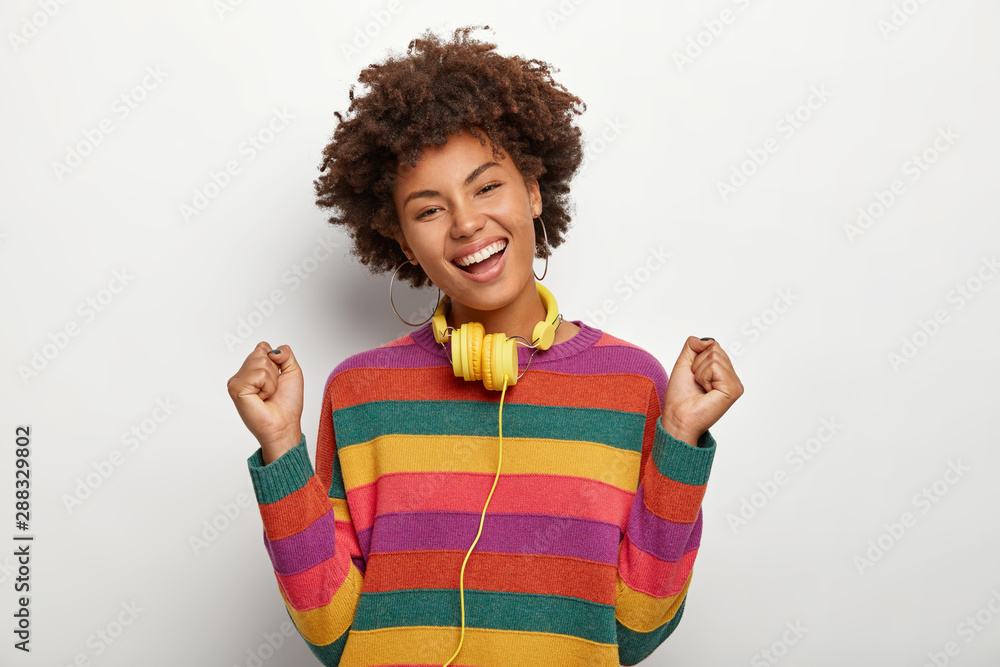 Upbeat dark skinned lady dances with joy, raises clenched fists, celebrates success and victory, wears colorful sweater, smiles broadly, poses over white background, has headphones for listening music Upbeat dark skinned lady dances with joy, raises clenched fists, celebrates success and victory, wears colorful sweater, smiles broadly, poses over white background, has headphones for listening music