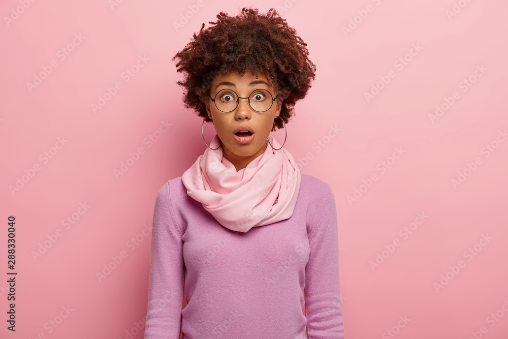 Emotional stupefied woman with curly Afro hairstyle, keeps mouth opened, wears round spectacles, casual jumper, shocked by puzzled news, opens mouth from wonder, isolated on pink background. Emotional stupefied woman with curly Afro hairstyle, keeps mouth opened, wears round spectacles, casual jumper, shocked by puzzled news, opens mouth from wonder, isolated on pink background.