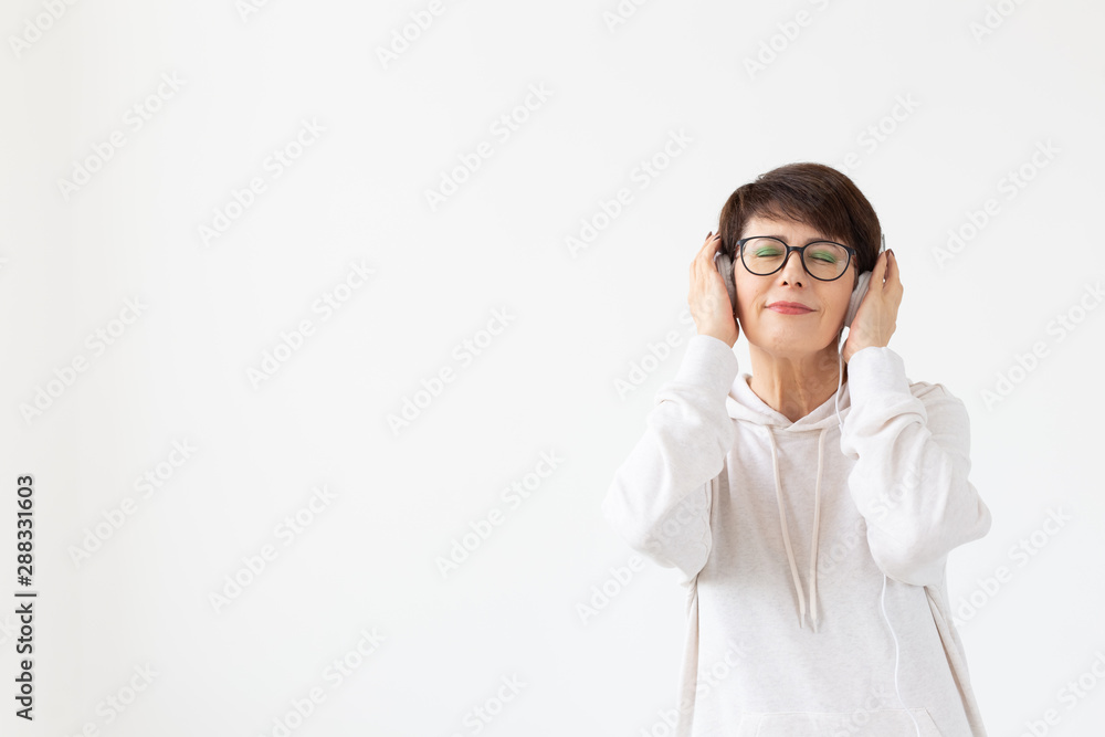 Pretty keen middle-aged woman in glasses and a white sweater listens to her favorite music with headphones on a white background with copy space. Online radio and music subscription concept. Pretty keen middle-aged woman in glasses and a white sweater listens to her favorite music with headphones on a white background with copy space. Online radio and music subscription concept.