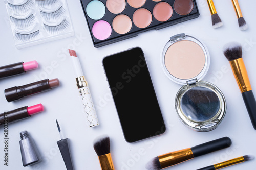 Creative arrangement of cosmetics on white background. Smartphone with blank display in the middle. 