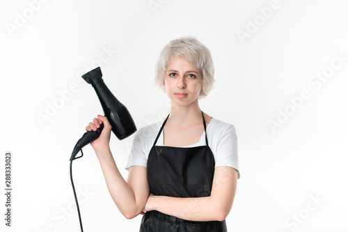 Young pretty hairdresser in white tee and black apron holding professional black hairdryer ready for work isolated on white background. Wellness and beauty industry and professional career.