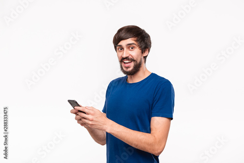 Handsome brunet man in a blue shirt with beard and mostaches surprised holding smartphone looking at the camera standing isolated over white background. Emotion and gesture of surprise.
