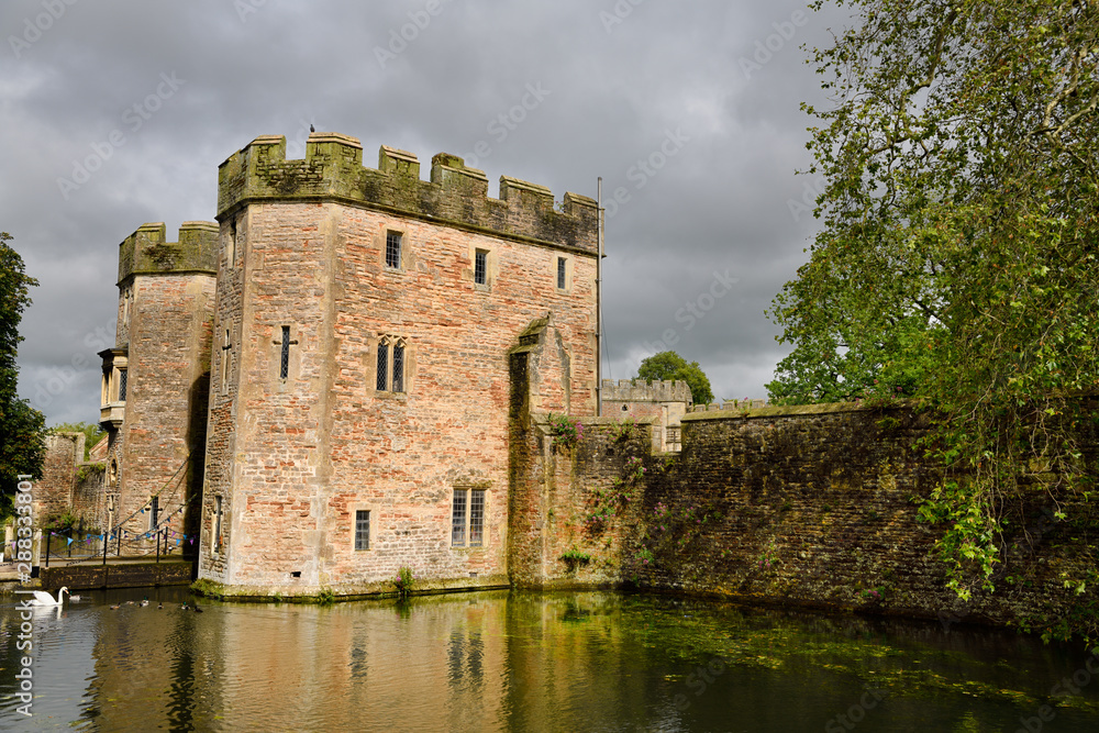 Sun on Bishop's Palace Gatehouse with bridge over the moat with swan and ducks under cloudy sky in Wells Somerset England