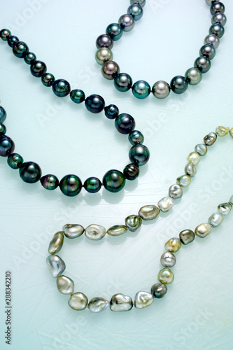 Three necklaces of single strand multi colored Tahitian pearls are laid on a frosted white surface.