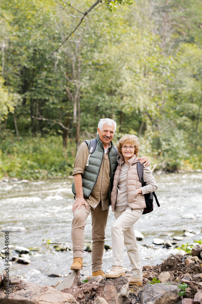 Happy aged active hikers with backpacks looking at you while standing by river Happy aged active hikers with backpacks looking at you while standing by river
