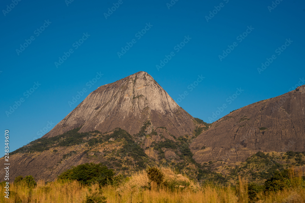 The rocky and conical shaped mountain rises steeply from the grassy plains under a clear blue sky in rural Mozambique, Africa