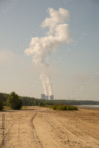 Cloud-sized steam rising from tall pipes visible behind a forest by the sea. Vertical orientation