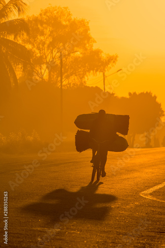 A backlit Mozambican cyclist with dramatic shadow, carrying a heavy load of coal to market along a tarmac road at sunrise. Nampula Town, Mozambique