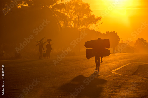 A Mozambican cyclist carrying large bags of coal to market at sunrise in the morning  along a tarmac road. Other commuters carrying items in the background. Nampula Town  Mozambique