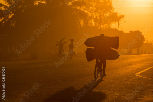 A Mozambican cyclist carrying large bags of coal to market at sunrise in the morning, along a tarmac road. Other commuters carrying items in the background. Nampula Town, Mozambique